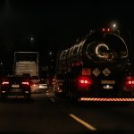 Veículos trafegam à noite por uma rodovia iluminada, com faróis e luzes destacadas na pista.