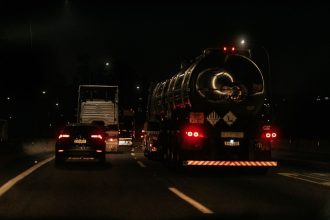 Veículos trafegam à noite por uma rodovia iluminada, com faróis e luzes destacadas na pista.