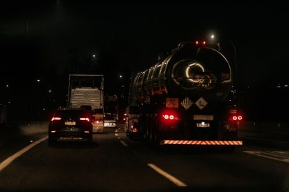Veículos trafegam à noite por uma rodovia iluminada, com faróis e luzes destacadas na pista.
