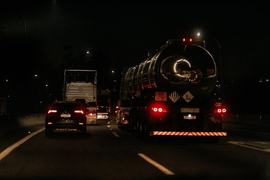 Veículos trafegam à noite por uma rodovia iluminada, com faróis e luzes destacadas na pista.