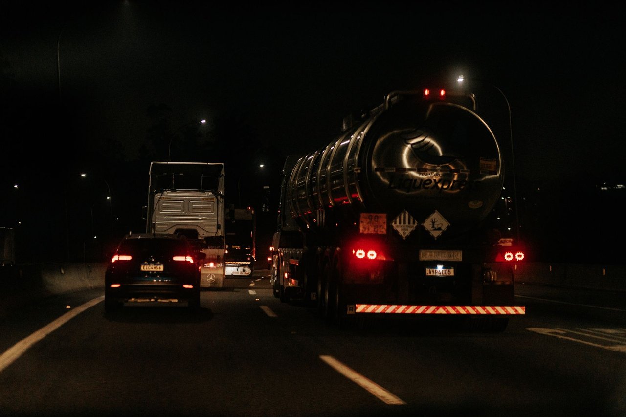 Veículos trafegam à noite por uma rodovia iluminada, com faróis e luzes destacadas na pista.