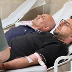 Dois homens doam sangue deitados em leitos hospitalares, acompanhados durante o procedimento de coleta.